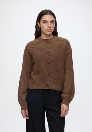 Kvinde med langt mørkt hår iført brun strikket cardigan med knapper og sorte bukser, står foran en ensfarvet lys baggrund.