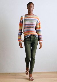 Multicolored gebreide trui met horizontale strepen, groene broek en witte sandaaltjes met bandjes. Model draagt een witte schoudertas.