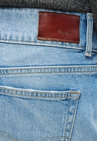 Vaqueros de mezclilla azul claro con una etiqueta de cuero marrón cosida en la cintura, con bolsillos traseros y detalle de costuras visibles.