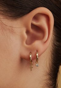 Goldene Creolen mit einem kleinen Kreuzanhänger und einem klaren Edelstein. Glatte, glänzende Oberfläche mit geschwungenen Formen, präsentiert an einem Ohr.