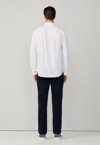 Camicia bianca a maniche lunghe con una piega sul retro, abbinata a pantaloni blu navy e sneakers blu. Linee pulite e design classico.
