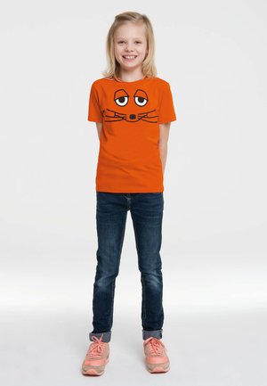 Orange t-shirt featuring a cartoon face with eyes, whiskers, and a nose. Paired with dark jeans and light pink sports shoes.