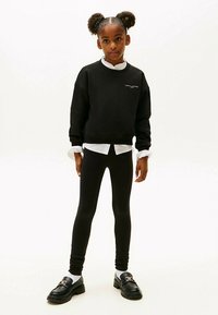 Sort sweatshirt med hvidt logo, lag på lag over en hvid skjorte med krave. Sorte leggings og sorte sko med spænde fuldender outfittet.