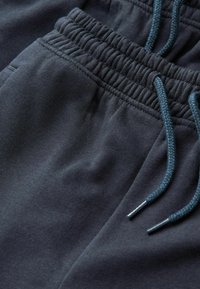 Dunkelgraue Jogginghose mit einem gerippten elastischen Bund, versehen mit einem türkisfarbenen Kordelzug und silbernen Metallenden. Glatte Stoffstruktur.