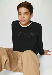 Schwarzes Langarmshirt mit dem Wort "CODE" in erhabenen Buchstaben, kombiniert mit lockeren, hellbeigen Hosen. Glatte Stofftextur.