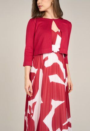 Rote, verkürzte Strickjacke mit Dreiviertelärmeln über einem plissierten Kleid mit einem auffälligen weißen abstrakten Muster auf rotem Hintergrund.