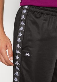 Černé sportovní kalhoty s elastickým pasem, na boku se pruhy s opakujícími se bílými logy a hladkým nylonovým povrchem.