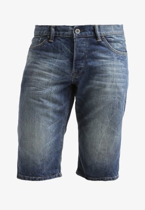 Czarny t-shirt z grafiką w stylu kreskówkowym, zestawiony z niebieskimi szortami dżinsowymi i czarnymi trampkami. Dopełnieniem stroju jest brązowy plecak.