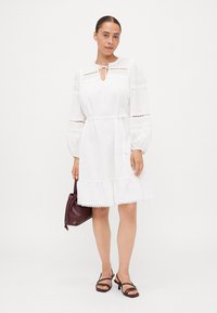 Vestido branco com mangas longas e bufantes, detalhes em renda e um laço no pescoço. Apresenta uma bainha rendilhada e é usado com uma mala escura e sandálias.