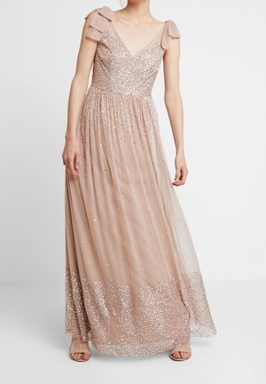 Kobieta ubrana w długą suknię do ziemi w kolorze różowym z cekinowymi zdobieniami, przezroczystą tiulową spódnicą i ramiączkami związanymi kokardami, stojąca na jednolitym tle.