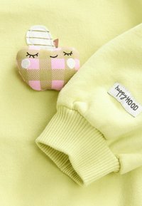 Gulgrön tyg med ribbade muddar har en texturerad, rutiga äpple-applikation i rosa och grönt, med broderade ansiktsdetaljer. Lappen säger "happyMOOD."