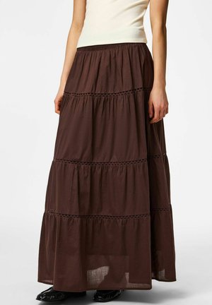 Mujer con una falda larga marrón de capas con ribete de encaje, combinada con una camiseta blanca sin mangas y zapatos negros, de pie frente a un fondo liso.