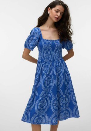 Kobieta w niebieskiej sukience midi z bufkami i białymi okrągłymi kwiatowymi wzorami, stojąca z rękami za plecami na jednolitym tle.