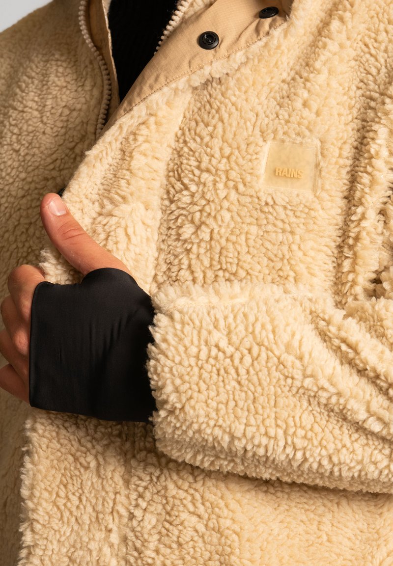 Giacca in pile color crema con una superficie morbida e texture. Presenta un colletto beige, polsini neri e un'etichetta con il logo sul davanti.