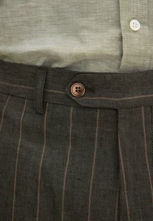 Gros plan sur un pantalon à fines rayures sombres avec une fermeture à bouton marron et une partie d'une chemise vert clair rentrée avec un bouton blanc.