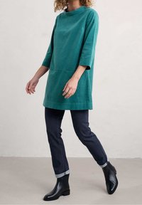 Miętowa tunika z wysokim dekoltem i rękawami do połowy przedramienia, z przednią kieszenią. Połączona z ciemnymi dżinsami i czarnymi krótkimi botkami.