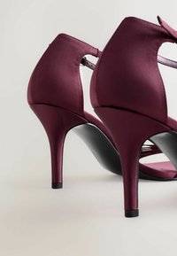 Zapatos de tacón alto de satén burdeos con un diseño elegante, parte posterior abierta y suela negra, con un tacón delgado y puntiaguado y tiras entrelazadas.