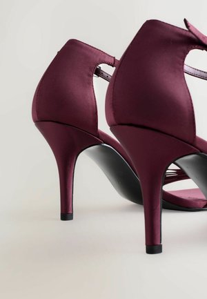 Zapatos de tacón alto de satén burdeos con un diseño elegante, parte posterior abierta y suela negra, con un tacón delgado y puntiaguado y tiras entrelazadas.