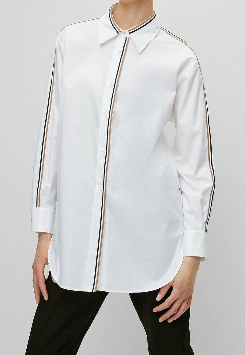 Bílá košile s dlouhým rukávem a límcem na knoflíky, s kontrastními černými a zlatými bočními pruhy a zakřiveným lemem.