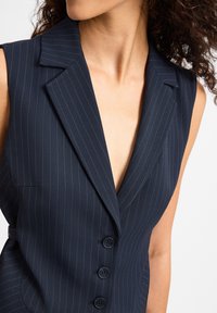 Marineblaues ärmelloses Blazer mit vertikalen Nadelstreifen, Reverskragen und fünf dunklen Knöpfen an der Vorderseite. Glatte Stoffstruktur.