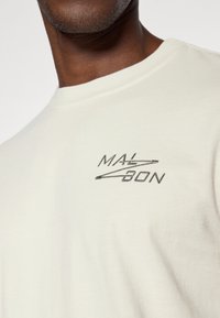 Ανοιχτό μπεζ βαμβακερό μπλουζάκι χαρακτηρισμένο από ένα διακριτικό μαύρο κεντητό λογότυπο που λέει "MAL-BON" με ένα σχεδιαστικό στοιχείο, στρογγυλή λαιμόκοψη και λείο υφή.