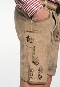 Beige lederhosen van Suède, met donkere borduursels, een voorzak en drie gouden knopen aan de zijkant. Zachte textuur en een slim-fit ontwerp.