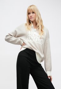 Világosszürke pulóver "Ski Club" felirattal, bordázott szegéllyel és mandzsettával, fekete nadrággal párosítva. Puha kötött anyag, laza szabás.