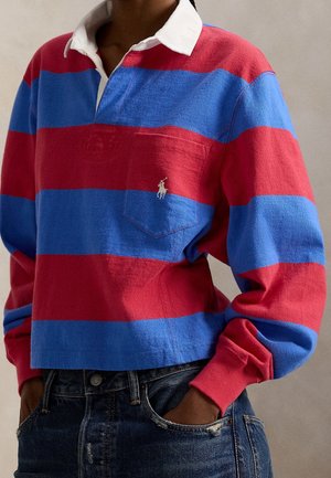 Personne portant un maillot de rugby à larges rayures rouges et bleues avec un col blanc et un logo sur la poche, associé à un jean en denim bleu foncé.