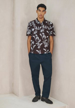 Dunkelbraunes kurzärmeliges Hemd mit weißem Blumenmuster, kombiniert mit dunkelblauer Hose und schwarzen Schuhen. Neutraler Hintergrund.