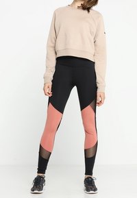 Sudadera corta beige con logo texturizado. Leggings negros con paneles geométricos rosas y detalles de malla. Zapatillas deportivas con diseño estampado.