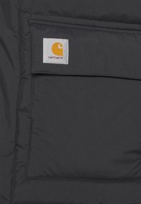 Acercamiento a un bolsillo de una chaqueta Carhartt negra con solapa cosida y parche de logo de la marca visible en amarillo y blanco.