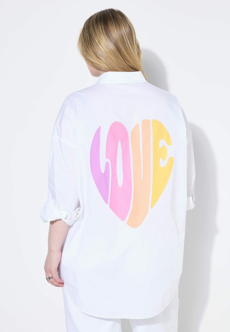 Hvid oversized skjorte med et farverigt hjerte-design i pink til gul, med ordet "KÆRLIGHED" i midten. Omslåede ærmer.