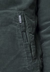 Donkergroene corduroy stof met verticale ribbels. Heeft een zijzak en een klein "Carhartt" label dat aan de rand is genaaid. Getextureerd oppervlak.