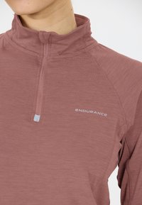 Close-up van een persoon die een mauve sporttop met kwart rits draagt, met het logo "ENDURANCE" op de borst.