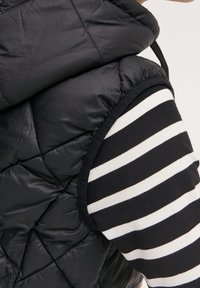 Musta tikattu liivi, jossa on korkea kaulus, yhdistetty mustavalkoraidalliseen pitkähihnaiseen paitaan. Kankaan tekstuuri ja kontrastiset kuviot.