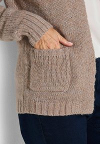 Hand ruht in der vorderen Tasche einer beigen gestrickten Strickjacke, die über einer dunklen Hose und einem hellen Hemd getragen wird.