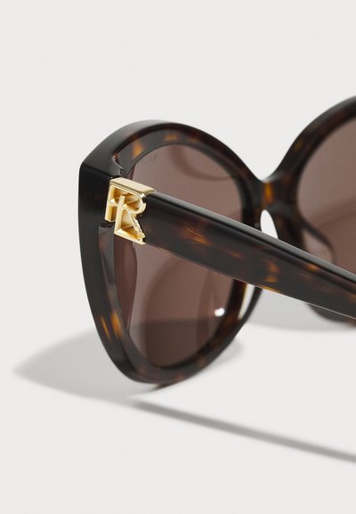 Brown tortoiseshell sunglasses with large, angular frames and a gold accent near the hinge. Lenses are dark brown, providing UV protection.