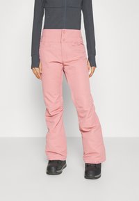 Rosa, isolierte Schneehosen mit schwingendem Schnitt, zwei Fronttaschen und Druckknopfverschluss. Getragen mit schwarzen Schneestiefeln und einem dunklen Oberteil.