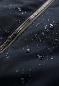 Tessuto impermeabile nero con una cerniera metallica visibile. Gocce d'acqua sparse sulla superficie evidenziano la sua texture resistente all'acqua.