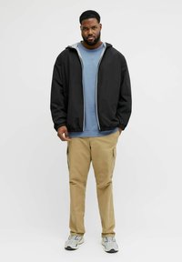 Sort jakke med glidelås og grå fôr, blå t-skjorte under, beige cargobukser og grå joggesko. Mann står foran en nøytral bakgrunn.