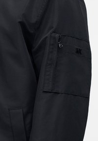 Μανίκι μαύρου μπουφάν με τσεπάκι με φερμουάρ και λεπτομέρεια λογότυπου "MK" κοντά στο ανώτερο μέρος του χεριού.