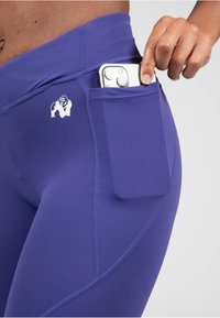 Paarse sportlegging met een zijzak voor een smartphone, zachte textuur en gestikte details, voorzien van een wit logo.