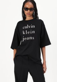 Černé oversized tričko s bílým nápisem "calvin klein jeans" v moderním písmu, s krátkými rukávy a volným střihem.