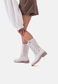 Stivaletti bianchi alla caviglia con motivi floreali intagliati al laser, materiale in pelle, tacco basso in legno e dettaglio hardware laterale. Presentano una finitura liscia.