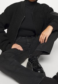 Black fuzzy bomber jacket with ribbed collar and cuffs, worn over a fitted black turtleneck. Paired with high-waisted black jeans and glossy shoes.