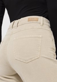 Beige corduroy pants with a tailored fit, featuring a single back pocket, button detail, and a leather label at the waist.