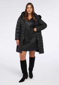 Abrigo acolchado negro con capucha de pelo sintético, cierre delantero con cremallera y detalles en dorado, llevado sobre un vestido negro de cuero y botas altas negras.