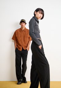Veste noire cintrée avec col haut et manches longues, associée à un pantalon noir taille haute. Chemise orange à manches courtes en arrière-plan.
