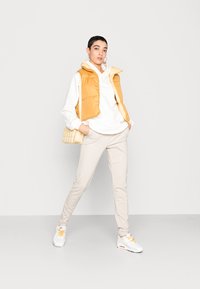 Colete amarelo acolchoado usado sobre um hoodie branco, combinado com calças bejas ajustadas e tênis brancos com detalhes em laranja. Pequena bolsa bege.