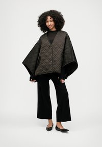 Casaco exterior preto matelassado em estilo poncho com decote em V, mangas largas e botões frontais, usado sobre uma blusa preta de mangas compridas e calças largas.